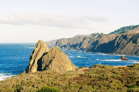 [The roadway winds atop short cliffs beside the ocean. The water is a beautiful blue some rocks in it. The hillsides below the road are dirt or rock, but the ones above the road do have trees.]